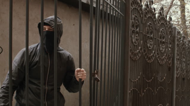 Bandit-man-in-black-mask-and-hood-with-baseball-bat-crawls-through-fence-mesh