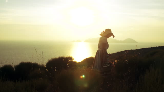 Schöne-junge-Frau-tragen-bunte-Kleid-Mode-mit-Rock-und-Hut-Kommissionierung-mit-Blumen-im-Korb-bei-Sonnenuntergang-auf-der-italienischen-Insel-Ponza-Berg-Küste.