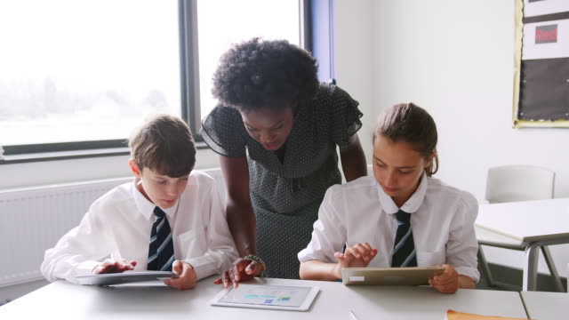 Weibliche-High-School-Lehrer-am-Tisch-mit-Studenten-in-Uniform-mit-digitalen-Tabletten-In-Lektion