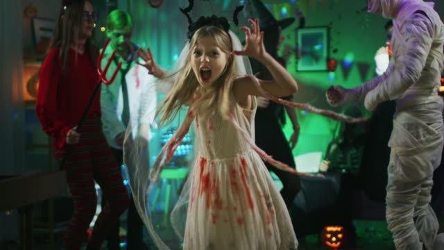 Halloween-Kostümparty:-Little-Girl-in-a-Bloody-White-Bride-Dress-dreht-sich-um-und-macht-Scary-Faces.-In-der-Hintergrundegruppe-der-Monster-tanzen-und-Spaß-haben-im-dekorierten-Raum-mit-Disco-Lights
