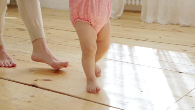 Tracking-shot-of-unrecognizable-cute-baby-girl-learning-to-walk-at-home-supported-by-her-loving-mother