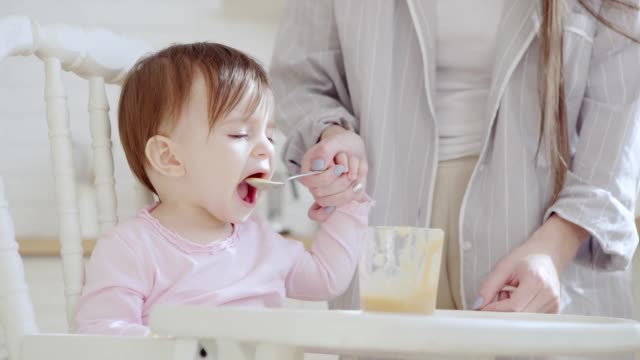 Unrecognizable-caring-mother-feeding-her-baby-daughter-fruit-puree-with-spoon.-Girl-refusing-to-eat.-Mom-wiping-her-hands-and-highchair-with-tissue