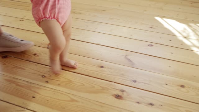 Spurensuche-von-nicht-erkennbarer-fürsorglicher-Mutter,-die-ihre-entzückende-Baby-Tochter-unterstützt,-die-sie-zu-Fuß-lernt-und-ihre-ersten-Schritte-auf-dem-Laubholzboden-zu-Hause-macht
