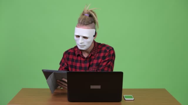 Hipster-man-wearing-mask-as-hacker-multitasking-work-against-wooden-table