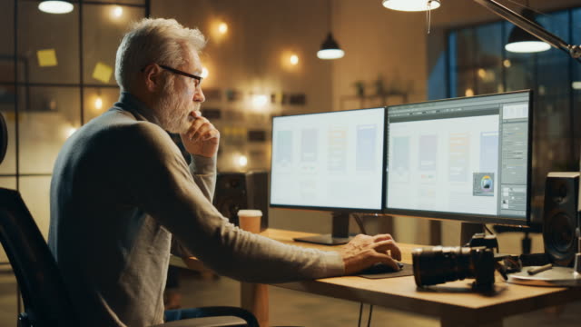 Creative-Middle-Aged-Mobile-Software-Designer-Sitting-at-His-Desk-Uses-Desktop-Computer-with-Two-Screens-Showing-Smartphone-Application-Design-Process.-Stylish-Modern-Studio-Office