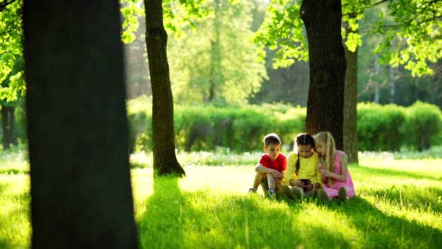 Foto-de-seguimiento-de-tres-niños,-dos-niñas-y-niño,-sentado-en-hierba-verde-bajo-el-árbol-en-el-parque-y-jugando-videojuegos-en-el-teléfono-inteligente