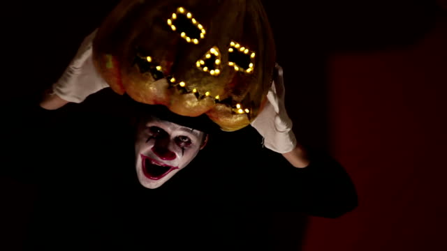 Gruseliger-Mann-im-Make-up-eines-Clowns-hält-einen-Kürbis-zu-Halloween-und-legt-ihn-auf-den-Kopf.-Schrecklicher-Clown-zieht-eine-Maske-in-Form-eines-Kürbisses-auf-den-Kopf.-Schrecklicher-Clown-hält-eine-Jack-O-Lantern-mit-leuchtenden-Augen-und-einem-Mu