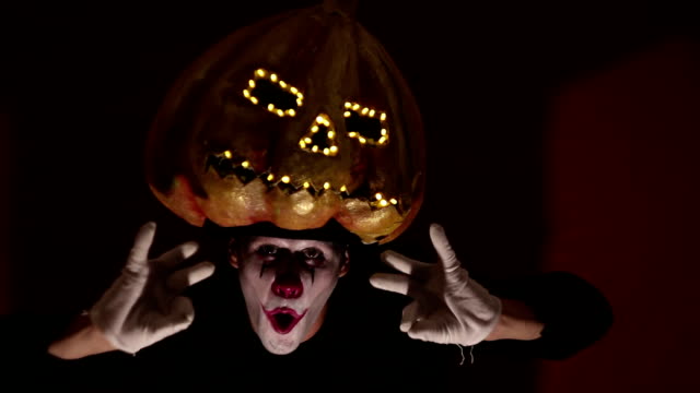 Gruseliger-Mann-im-Make-up-eines-Clowns-hält-einen-Kürbis-zu-Halloween-und-legt-ihn-auf-den-Kopf.-Schrecklicher-Clown-zieht-eine-Maske-in-Form-eines-Kürbisses-auf-den-Kopf.-Schrecklicher-Clown-hält-eine-Jack-O-Lantern-mit-leuchtenden-Augen-und-einem-Mu