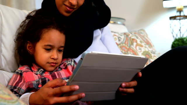 Joven-madre-usando-una-tableta-digital-con-su-hija-en-el-sofá-en-casa-4k