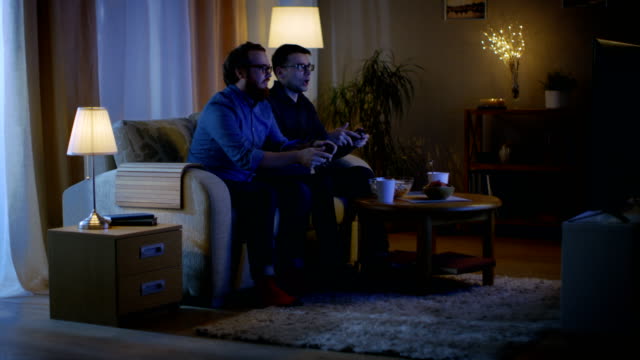 In-the-Evening-sitzen-zwei-Freunde-auf-einem-Sofa-im-Wohnzimmer-und-spielen-wettbewerbsfähige-Videospiele.-Einer-von-ihnen-gewinnt-und-er-genießt-seinen-Erfolg.