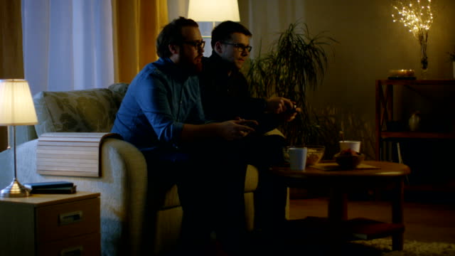 En-la-noche-dos-amigos-están-sentados-en-un-sofá-en-la-sala-de-estar-y-jugando-juegos-de-video-competitivos.