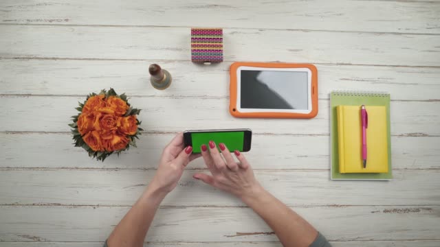 Frau-nimmt-eine-Smartphone-von-einem-weißen-hölzernen-Schreibtisch-und-tippen-mit-ca.-in-der-Nähe-sind-Blumen-und-eine-orange-digital-Tablette.-Chroma-Key-Green-Screen.-Ansicht-von-oben.-Hände,-Nahaufnahme