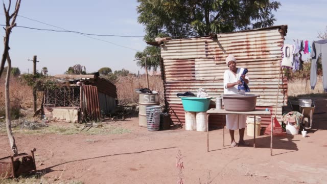 Mujer-africana-sin-agua-lavando-la-ropa-en-un-cubo-delante-de-su-choza-de-lata-casera