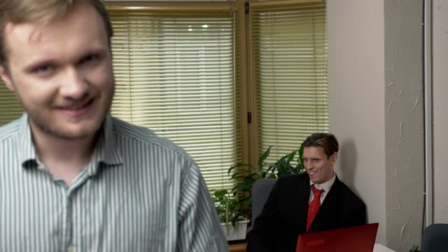 Young-man-with-a-beard-speaks-and-looks-at-the-camera,-a-guy-in-a-suit-in-the-background-makes-funny-faces,-fool-around.-Work-in-the-office-concept.-60-fps