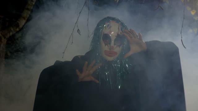 Malefic-halloween-funny-clown-wearing-a-mask-and-green-fake-hair-performing-an-evil-dance-in-a-dark-forest