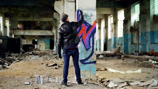 Vista-posterior-de-pintor-de-graffiti,-creando-la-bella-imagen-con-pintura-en-aerosol-dentro-de-edificio-abandonado.-Artista-está-usando-blue-jeans,-chaqueta-de-cuero-negro,-gorro-y-guantes.