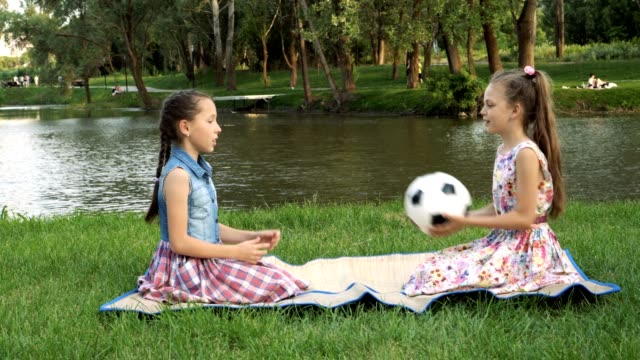 Two-charming-little-girls-sit-at-sunset-in-the-Park-on-the-lawn-against-the-river-and-play-with-a-soccer-ball,-throwing-it-to-each-other.