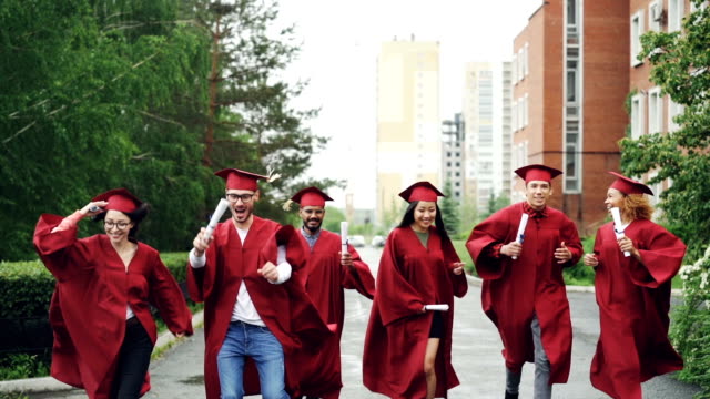 Emocionados-a-los-estudiantes-graduados-que-corre-a-lo-largo-de-la-carretera-en-el-campus-con-diplomas-usando-sombreros-y-vestidos-de-ropa-de-la-graduación.-Pequeña-lluvia-es-visible