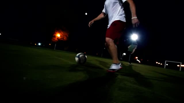 Young-man-in-Burgundy-shorts-and-white-sneakers-leads-the-ball-on-the-green-field,-night-shooting