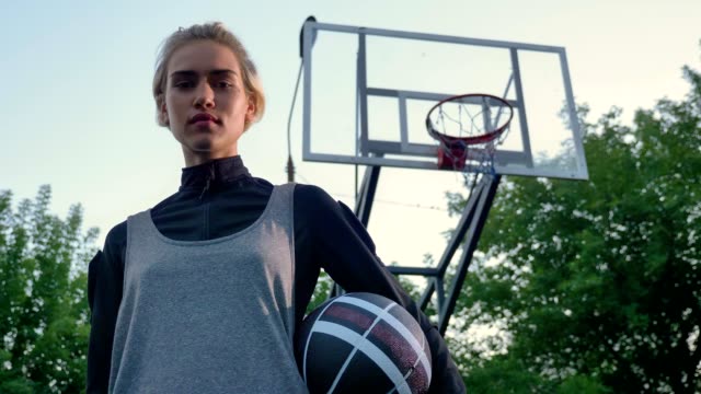 Beautiful-young-female-basketball-player-holding-ball-and-looking-at-camera,-standing-in-park,-low-angle