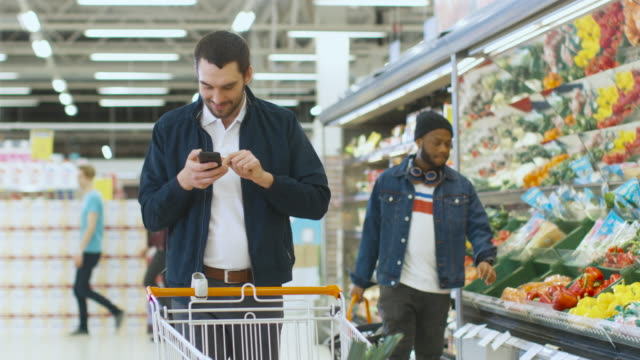 Im-Supermarkt:-gut-aussehender-Mann-mit-Smartphone,-lächelt-Spaziergänge-durch-frische-produzieren-Abschnitt-des-Speichers,-holt-Gemüse-und-Durchführungen-in-Warenkorb.