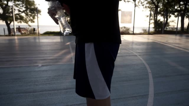 Down-up-footage-of-a-young-sporty-girl-basketball-player-have-a-rest-after-training-on-the-local-outdoors-court,-drinking-a-water-from-a-plastic-bottle