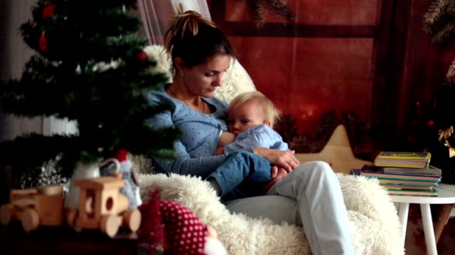 Mother-breastfeeding-her-toddler-son-sitting-in-cozy-armchair-near-Christmas-tree,-wintertime