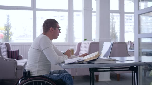 moderne-Technologien-in-der-Bildung,-Student-aus-Online-Unterricht-und-Notizen-im-Notizbuch-am-Tisch-im-Café-sitzen-lernen-Behinderte-im-Rollstuhl-mit-Laptop-arbeiten