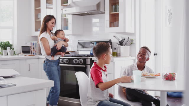 Mother-holding-baby-stands-in-the-kitchen-talking-with-her-son-and-his-friend,-over-for-a-playdate