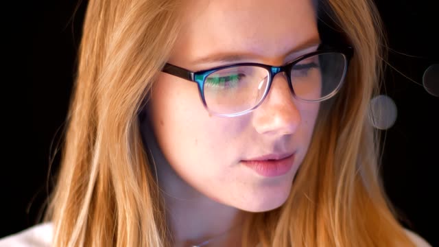 Concentrated-face-close-up,-blonde-in-spectacles-is-looking-at-phone-screen-while-standing-,-green-screen-reflected-in-glasses,-dark-background,-smart-person