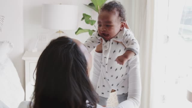 Millennial-madre-raza-mixta-levantando-a-su-bebé-y-sonriendo-a-él,-sobre-el-hombro-vista