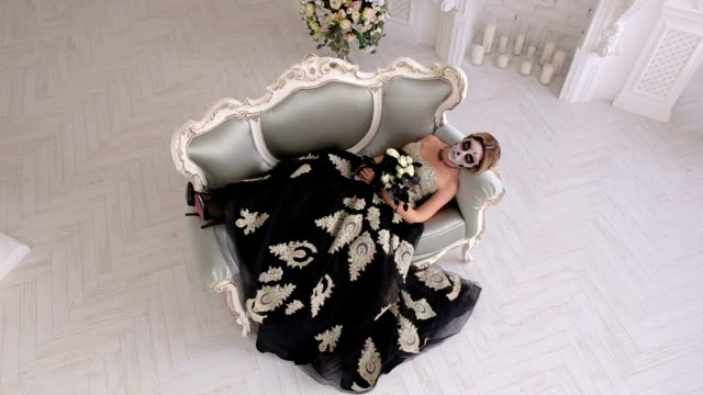 A-creepy-woman-in-a-dress-and-with-makeup-in-form-of-a-skull-lies-on-retro-sofa