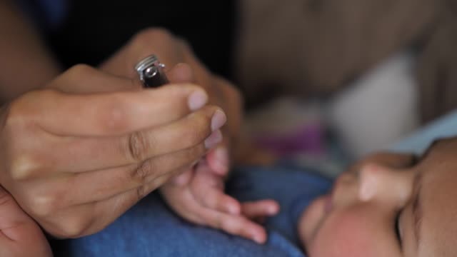 A-close-up-of-a-mixed-race-African-American-mother-clipping-one-of-her-baby's-fingernails-as-he-watches-as-he-lays-on-his-back-on-the-bed.