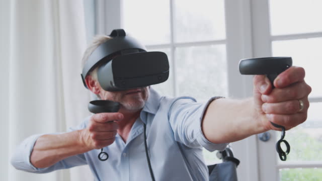 Senior-Disabled-Man-In-Wheelchair-At-Home-Wearing-Virtual-Reality-Headset-Holding-Gaming-Controllers