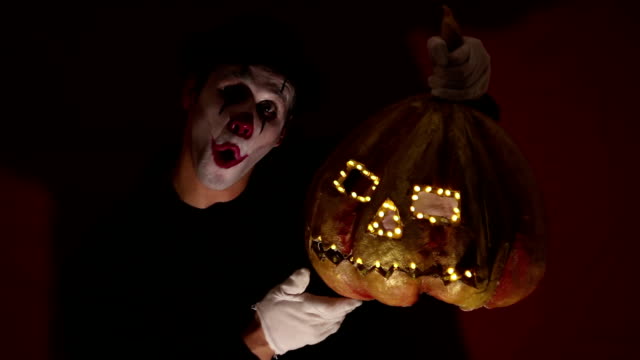 Scary-clown-holds-a-pumpkin-mask-in-his-hands.-A-scary-man-in-a-clown-makeup-holds-a-pumpkin-for-Halloween.-A-scary-clown-holds-a-Jack-O-Lantern-in-his-hands-with-luminous-eyes-and-a-mouth.