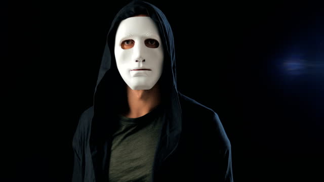 Anonymous-man-in-a-white-mask-covering-his-face-and-a-black-hood-turns-and-looks-at-the-camera.-Black-dark-background.-Concept-of-a-member-of-a-secret-organization