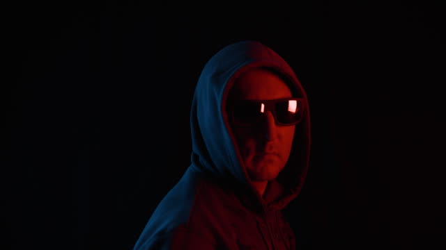 Adult-man-in-black-hood-and-sunglasses-turning-and-looking-to-camera-on-black-background.-Unrecognizable-man-in-black-hoodie-and-glasses-in-blue-and-red-lighting-in-dark-studio.