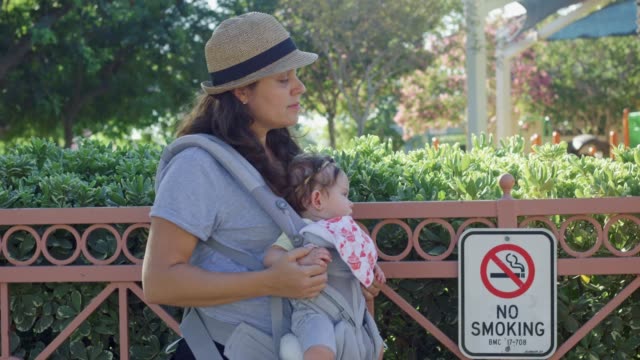 Mujer-y-bebé-en-el-portador-de-pie-junto-a-no-fumar-cartel-en-el-parque