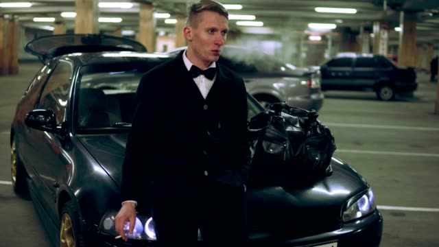 Young-man-in-a-black-suit-with-a-bow-tie-smoking-a-cigarette-sitting-at-the-bonnet-of-the-black-car-in-the-parking.-Waiting-for-someone.-Front-view.