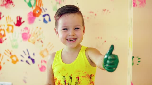 Close-up-veiw-of-cute-little-boy-with-painted-hands-after-leaving-his-colorful-handprints-on-the-wall-and-showing-his-thumb-up.-Young-happy-family.-Mother-and-child-concept