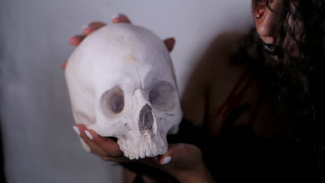 Scary-portrait-of-young-girl-with-Halloween-blood-makeup-holding-real-skull.-Beautiful-latin-woman-with-curly-hair-posing-in-studio.-Slow-motion