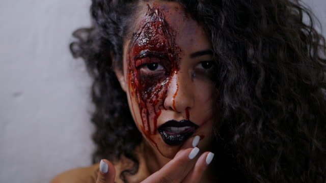 Scary-portrait-of-young-girl-with-Halloween-makeup-smears-blood-on-her-face.-Beautiful-latin-woman-with-curly-hair-looking-into-camera-in-studio.-Slow-motion
