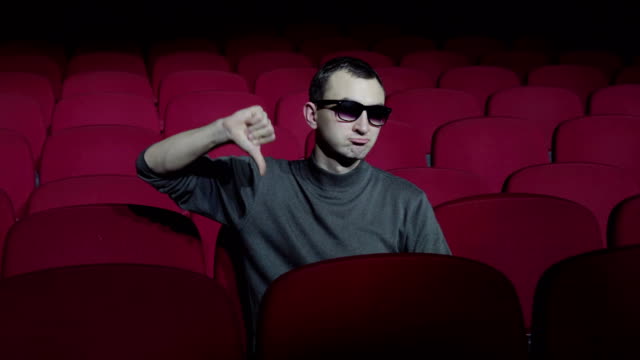 Single-man-sitting-in-comfortable-red-chairs-in-dark-cinema-theater-and-showing-thumbs-down