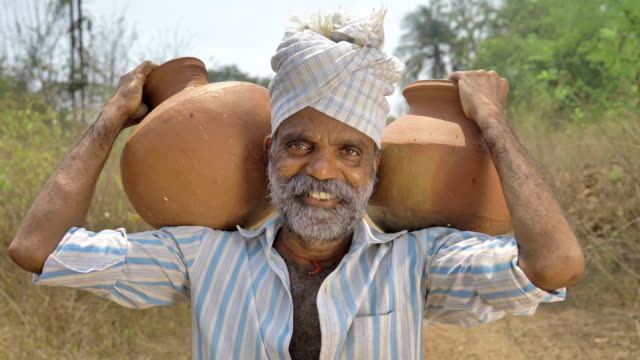 Un-agricultor-hombre-feliz-y-sonriente-con-dos-ollas-de-barro-lleno-de-agua-sobre-sus-hombros-en-campo.