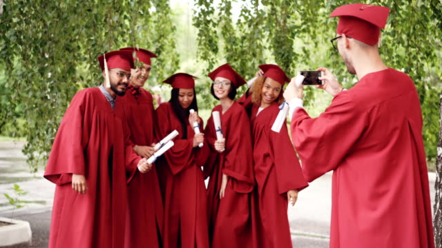 Fröhliche-Absolventen-stehen-in-der-Schlange-im-freien-und-junger-Mann-mit-Smartphone-ist-erschießen-während-Absolventen-lachen,-posieren,-Hände-winken.