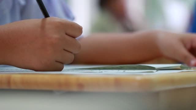 Education-test-concept-:-Hands-student-holding-pen-for-testing-exams-writing-answer-sheet-or-exercise-for-taking-fill-in-exam-carbon-paper-computer-on-wood-table-at-classroom-in-high-school