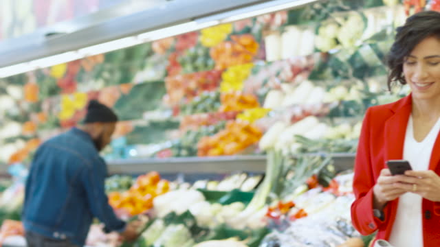 At-the-Supermarket:-Beautiful-Young-Woman-Uses-Smartphone-While-Standing-at-the-Fresh-Produce-Section-of-the-Store.-In-the-Background-Other-Customers-Shopping.