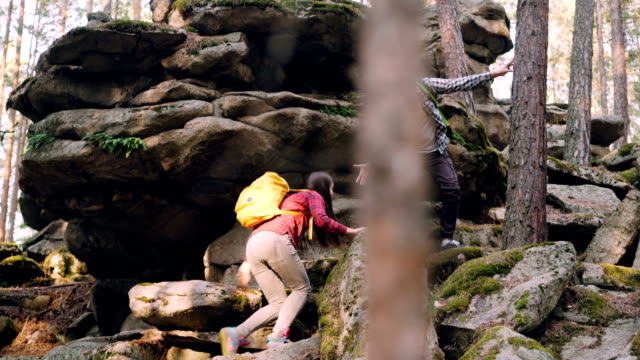 Adventurous-young-people-friends-are-trekking-in-forest-climbing-up-huge-rocks,-guy-is-helping-girl-giving-her-hand-during-hard-walk.-Adventures-and-friends-concept.