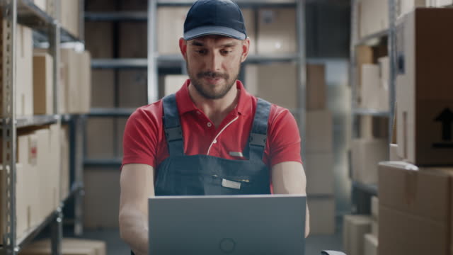 Worker-Uses-Laptop-while-Sitting-at-His-Desk-in-the-Warehouse.