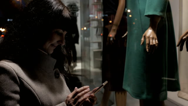 Shopping-at-the-mall,-sales-time.-Woman-looking-at-boutique-showcase-in-the-evening-city-on-the-street.-Beautiful-attractive-woman-near-shop-window-with-mannequins.-Reading-bar-codes-by-smartphone.